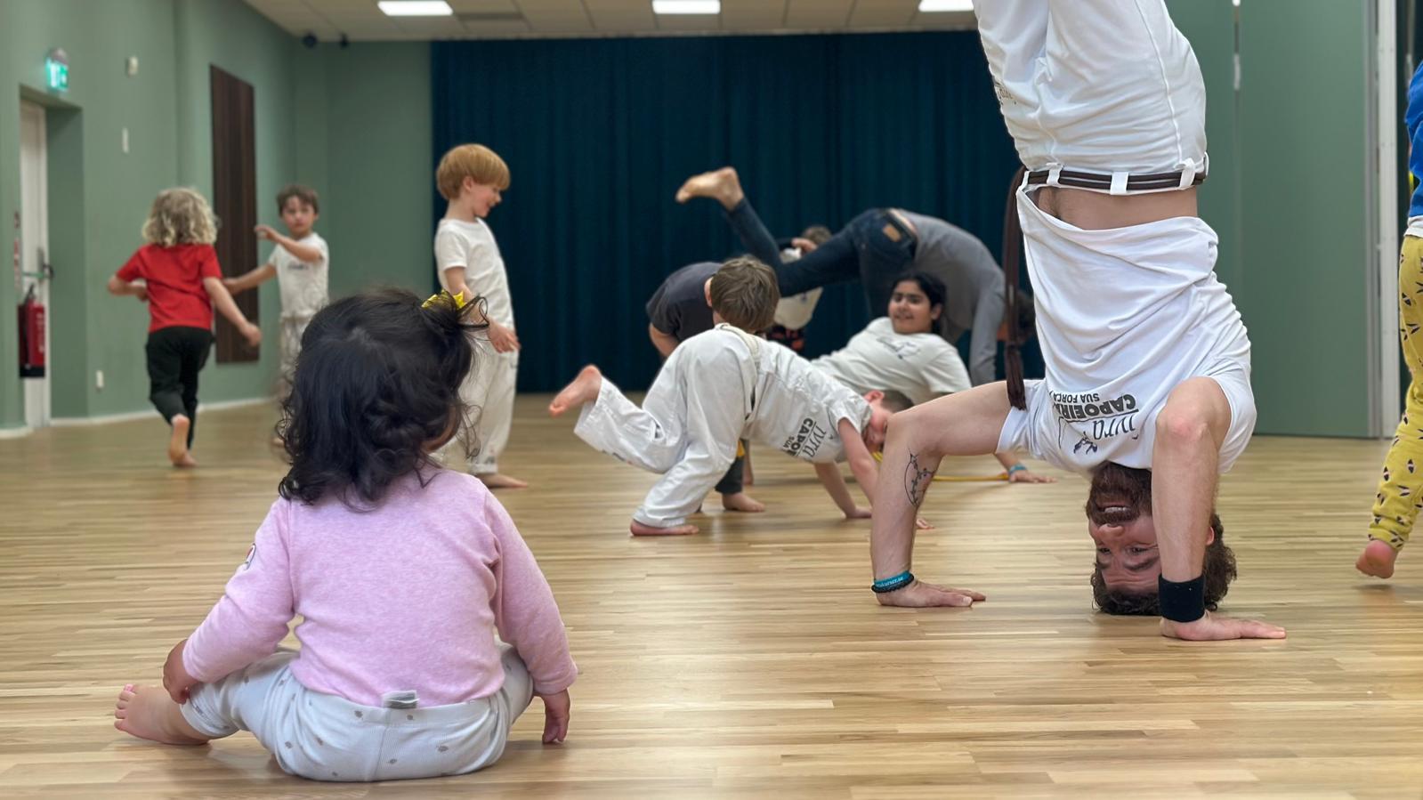 Capoeiraträning hos Ivra: barn och vuxna tränar kampsport, akrobatik och musik tillsammans i ljus träningslokal.
